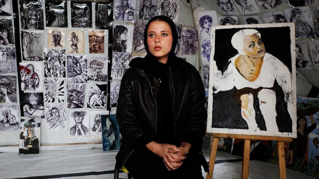 الفنانة الفلسطينية الشابة، مرح خالد تجلس أمام إحدى لوحاتها في الخيمة التي حولتها إلى معرض فني في قلب مدرسة تابعة لوكالة الأونروا التي تحولت إلى مركز لإيواء النازحين في مدينة غزة.